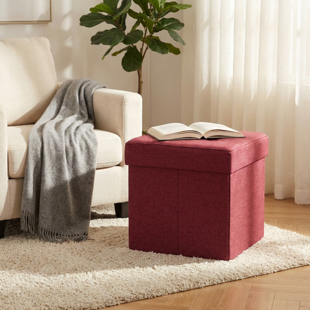 Pouf contenitore quadrato in tessuto bordeaux ambientato in un living luminoso accanto a una poltrona chiara.