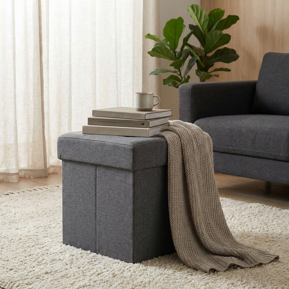 Pouf quadrato grigio scuro utilizzato come piano d'appoggio per libri e tazza accanto a un divano.