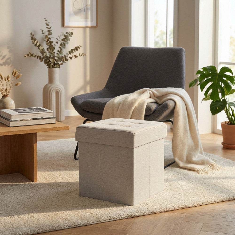 Pouf contenitore quadrato in tessuto tortora ambientato in un salotto luminoso accanto a una poltrona grigia.
