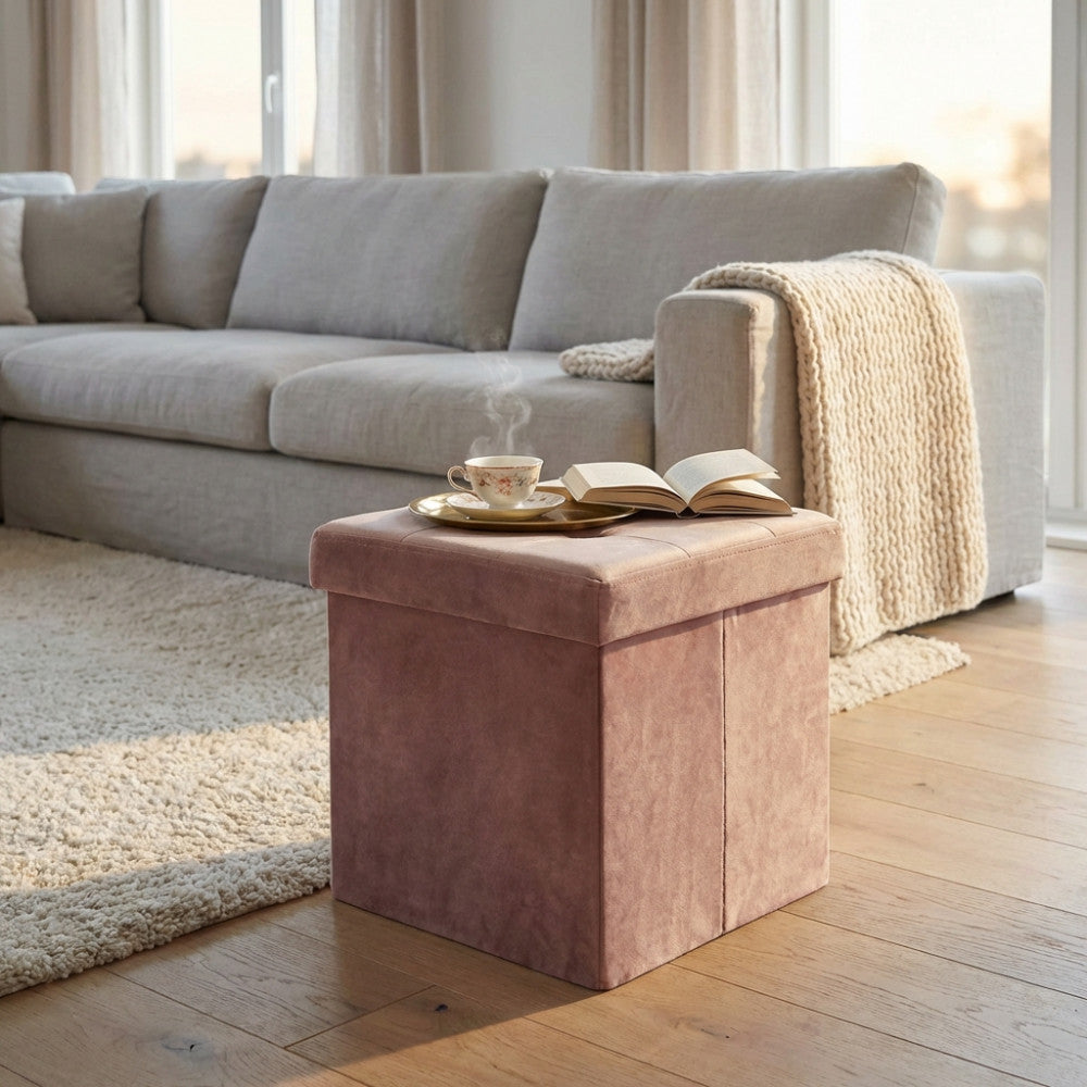 Pouf quadrato in velluto rosa nel soggiorno, accanto a un divano grigio, con una tazza di tè fumante e un libro aperto.