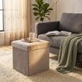 Pouf contenitore quadrato in velluto tortora ambientato in un salotto luminoso accanto a un divano grigio.