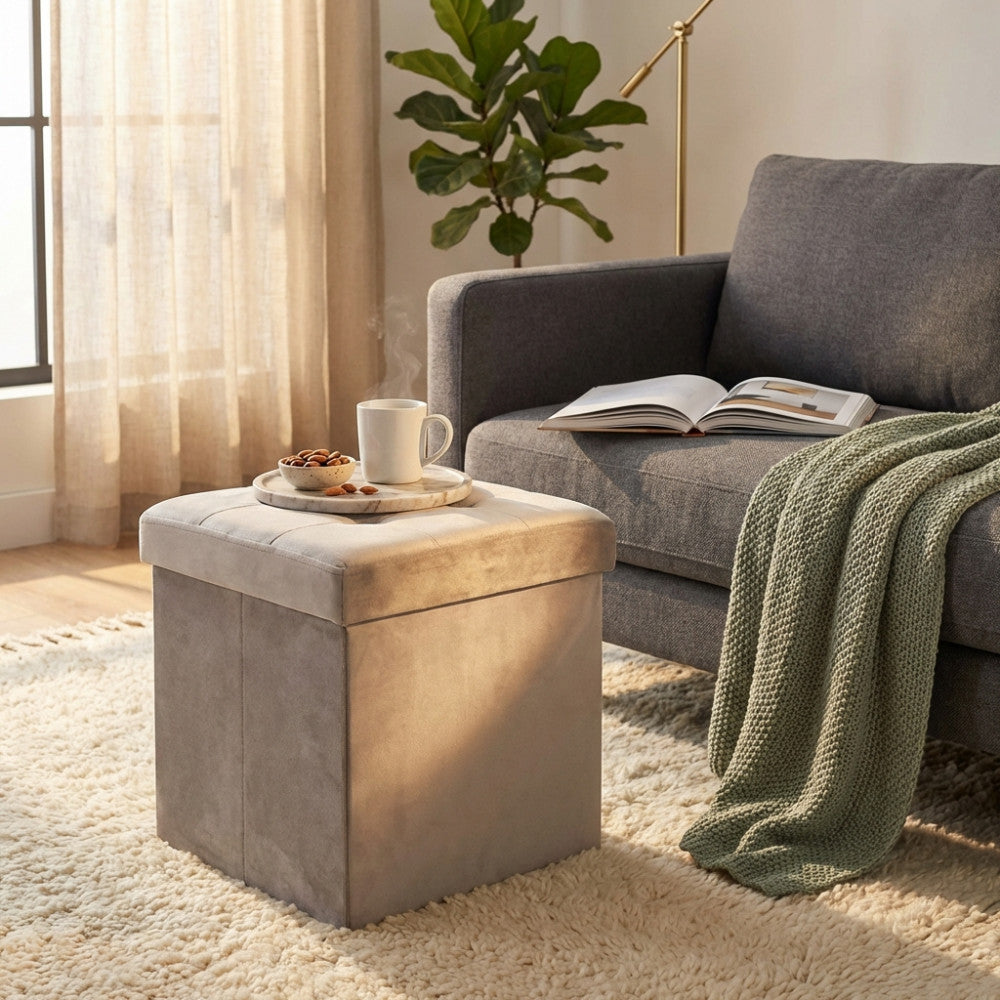 Pouf contenitore quadrato in velluto tortora ambientato in un salotto luminoso accanto a un divano grigio.
