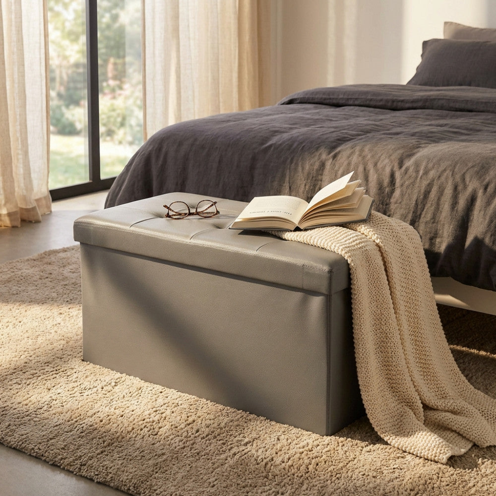 Pouf contenitore rettangolare in ecopelle grigia posizionato ai piedi di un letto matrimoniale, con un libro aperto e occhiali appoggiati sopra.