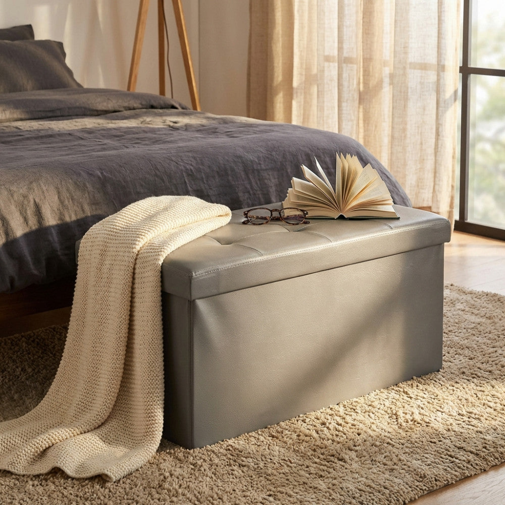 Dettaglio del pouf contenitore in ecopelle grigia in una camera da letto luminosa, con una morbida coperta in maglia adagiata sulla seduta.