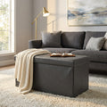 Pouf contenitore rettangolare in ecopelle nera ambientato in un salotto luminoso, decorato con una coperta in maglia e un libro aperto.
