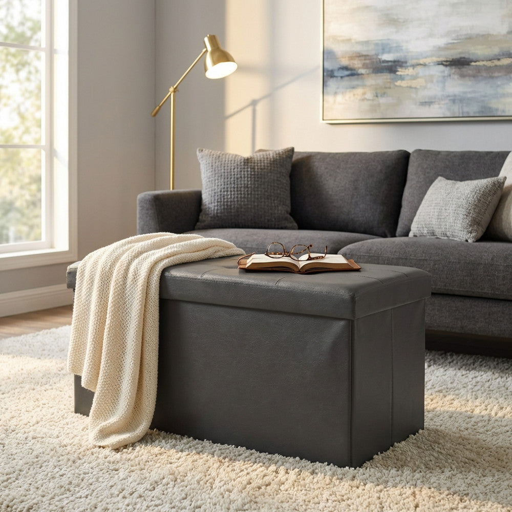 Pouf contenitore rettangolare in ecopelle nera ambientato in un salotto luminoso, decorato con una coperta in maglia e un libro aperto.