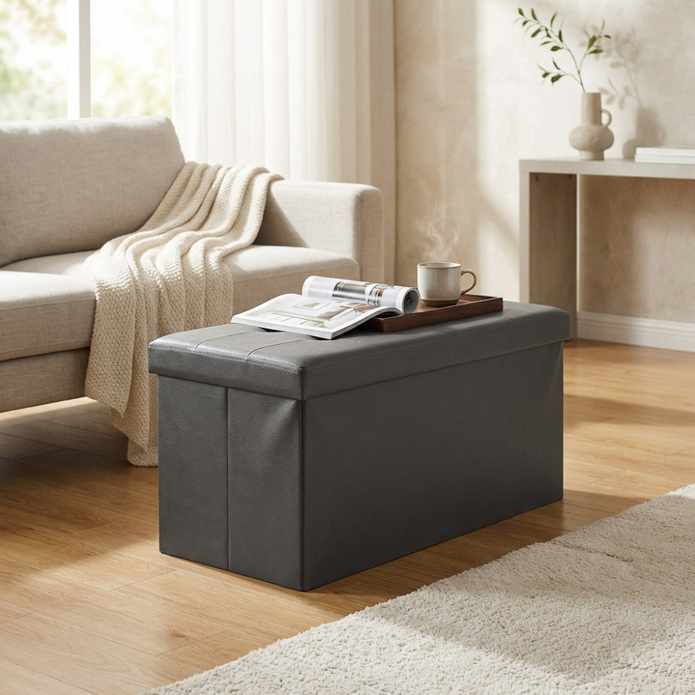 Pouf in ecopelle nera utilizzato come tavolino da salotto, con un vassoio in legno, una tazza fumante e una rivista.