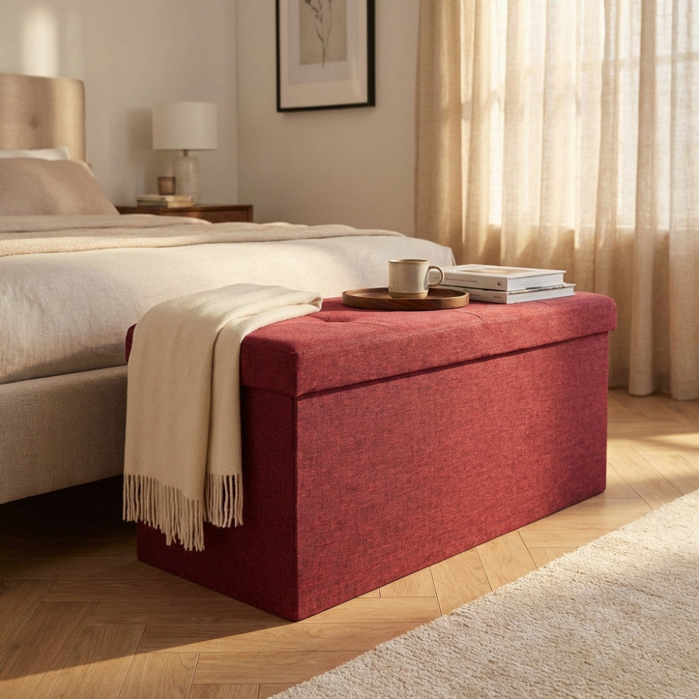 Pouf contenitore rettangolare in tessuto bordeaux posizionato ai piedi di un letto matrimoniale in una camera luminosa.
