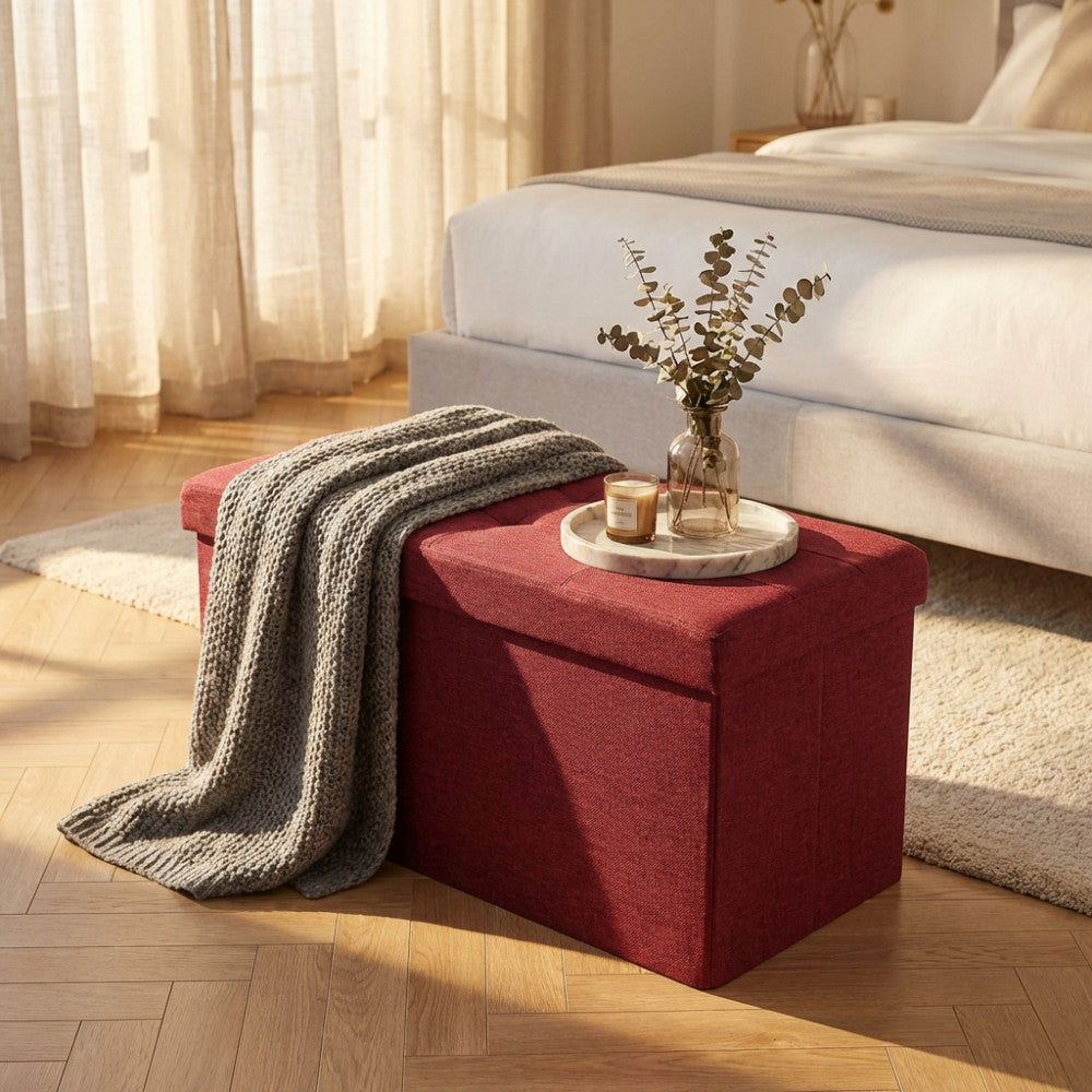 Pouf bordeaux allestito con una coperta in maglia grigia, un vassoio in marmo, una candela e un vaso con rami decorativi.