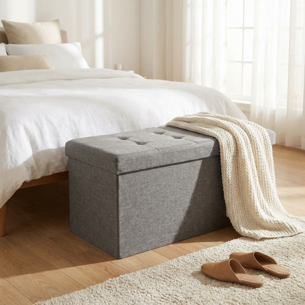 Pouf contenitore rettangolare in tessuto grigio posizionato ai piedi di un letto in una camera luminosa.