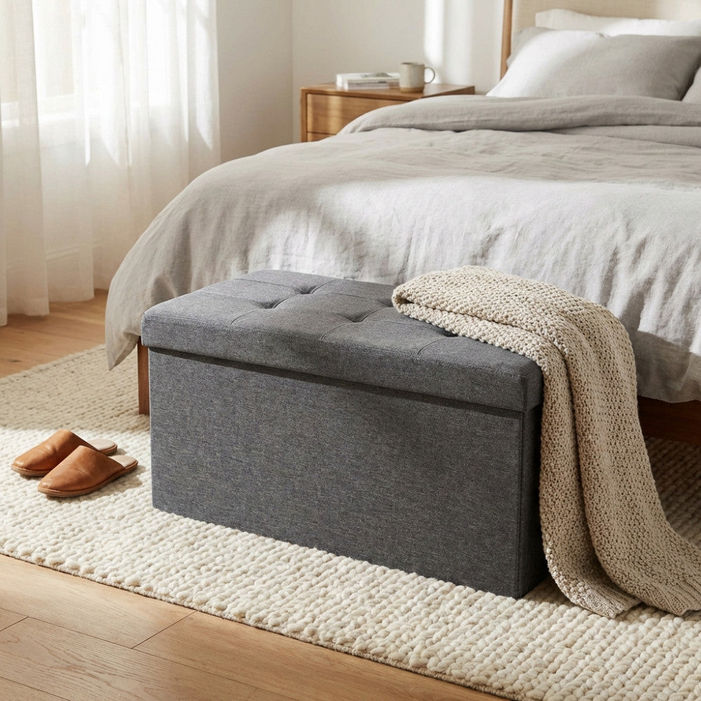 Pouf contenitore rettangolare in tessuto grigio scuro posizionato ai piedi di un letto matrimoniale, con una coperta in maglia appoggiata sopra.