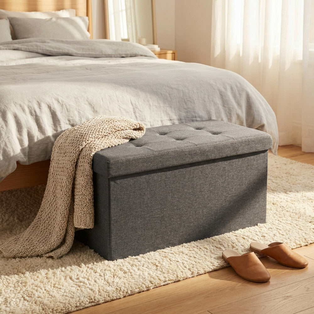 Dettaglio del pouf contenitore grigio scuro in un ambiente camera da letto luminoso, che evidenzia la texture del tessuto e la seduta trapuntata.
