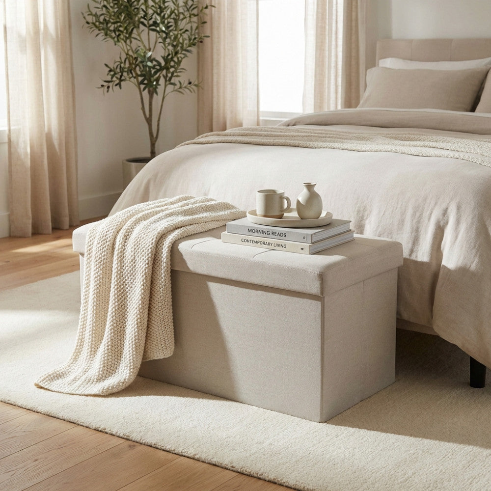 Dettaglio del pouf contenitore in tessuto tortora con libri, una tazza e una coperta in maglia appoggiati sopra.