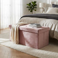 Pouf contenitore rettangolare in velluto rosa posizionato ai piedi di un letto matrimoniale, con una coperta chiara e libri appoggiati sopra.