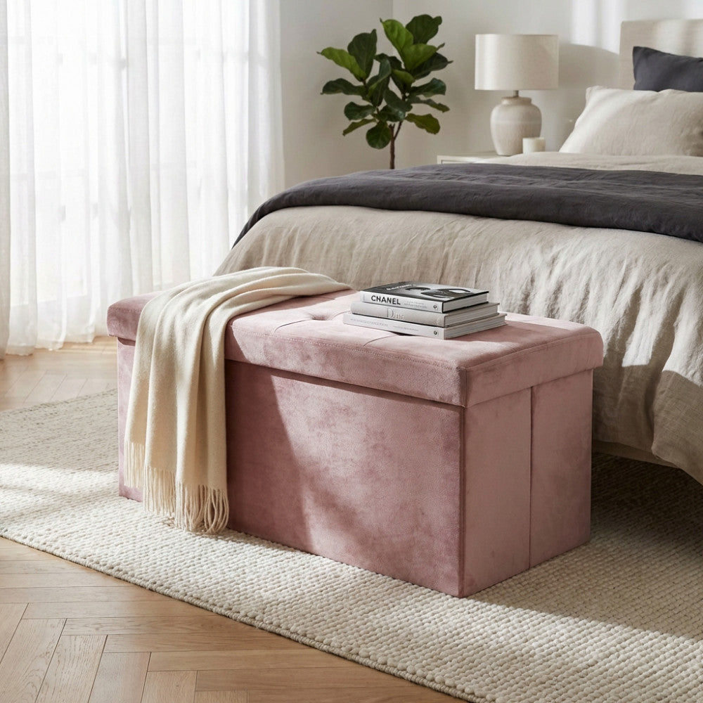 Pouf contenitore rettangolare in velluto rosa posizionato ai piedi di un letto matrimoniale, con una coperta chiara e libri appoggiati sopra.