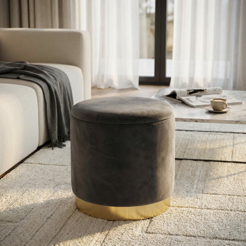 Pouf contenitore rotondo in velluto grigio scuro con base dorata, ambientato in un living luminoso accanto a un divano chiaro.