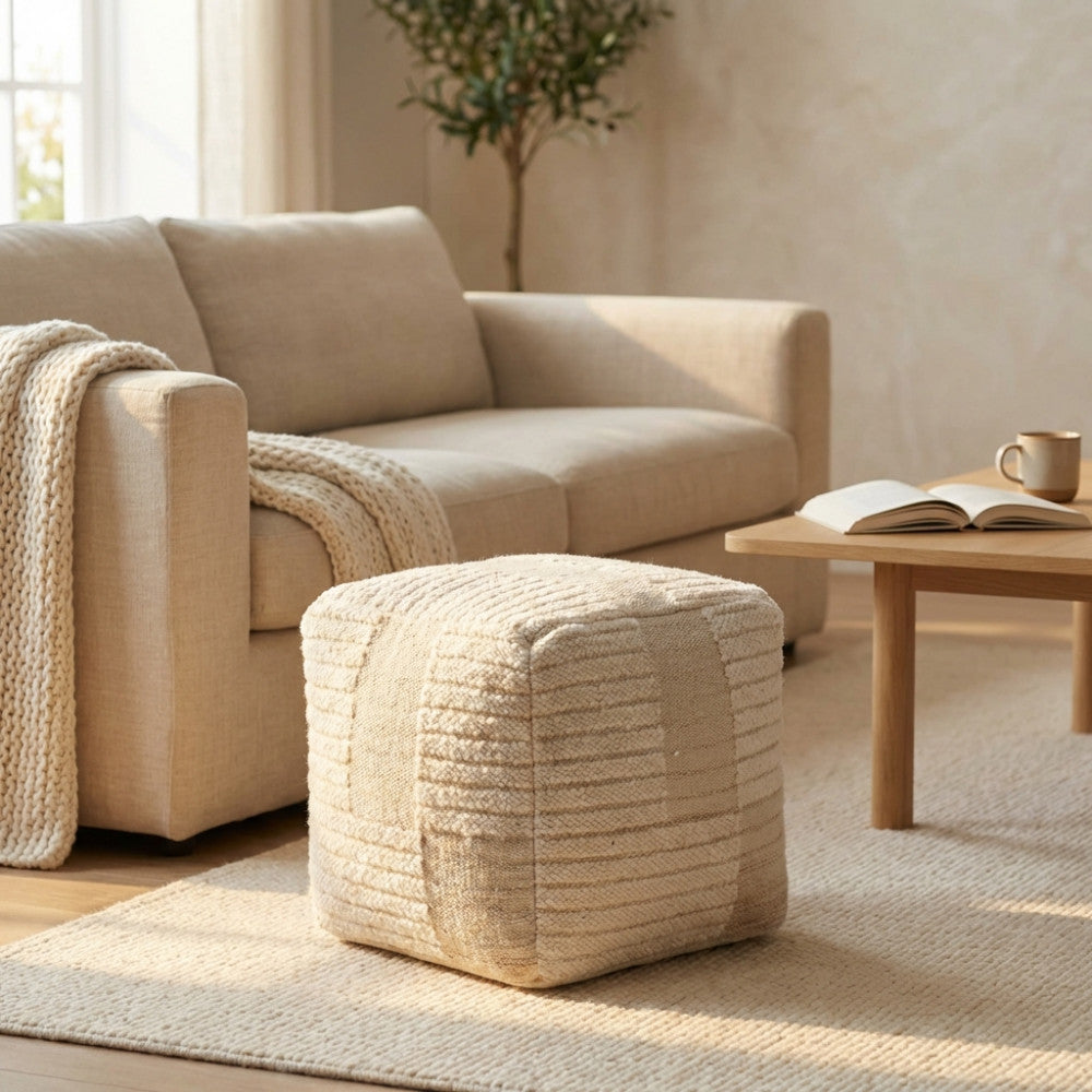 Pouf Josselin in tessuto bianco ecrù posizionato su un tappeto chiaro in un salotto luminoso, accanto a un divano beige.