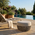 Pouf Lafayette beige in corda e alluminio posizionato a bordo piscina accanto a un lettino prendisole.