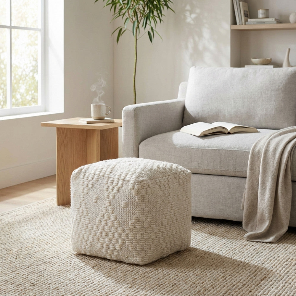 Pouf Marcel in lana e cotone bianco posizionato accanto a un tavolino in rovere chiaro e un divano grigio moderno in un soggiorno luminoso.