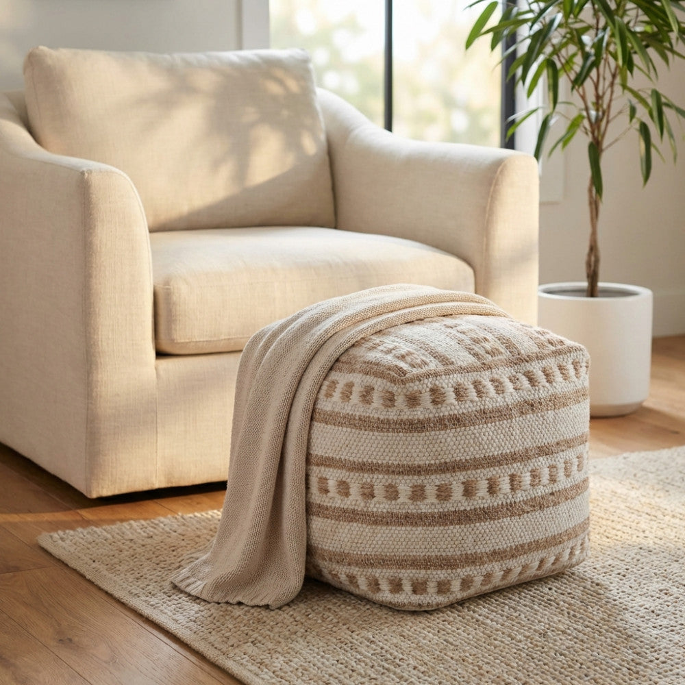 Pouf moderno materico decorato con una coperta in maglia beige, posizionato su un tappeto vicino a una poltrona moderna e una pianta in vaso.