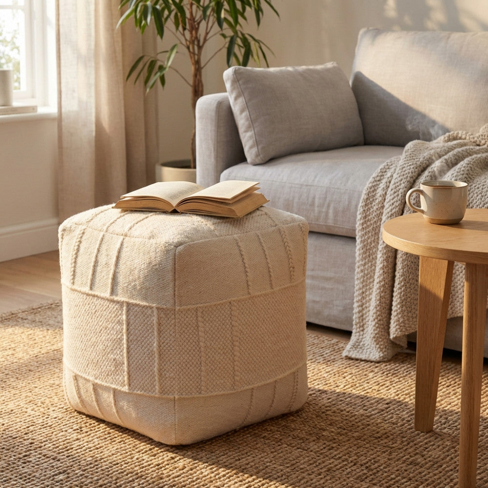 Pouf quadrato in lana e cotone bianco posizionato su un tappeto in iuta in un salotto luminoso, con un libro aperto sulla seduta.