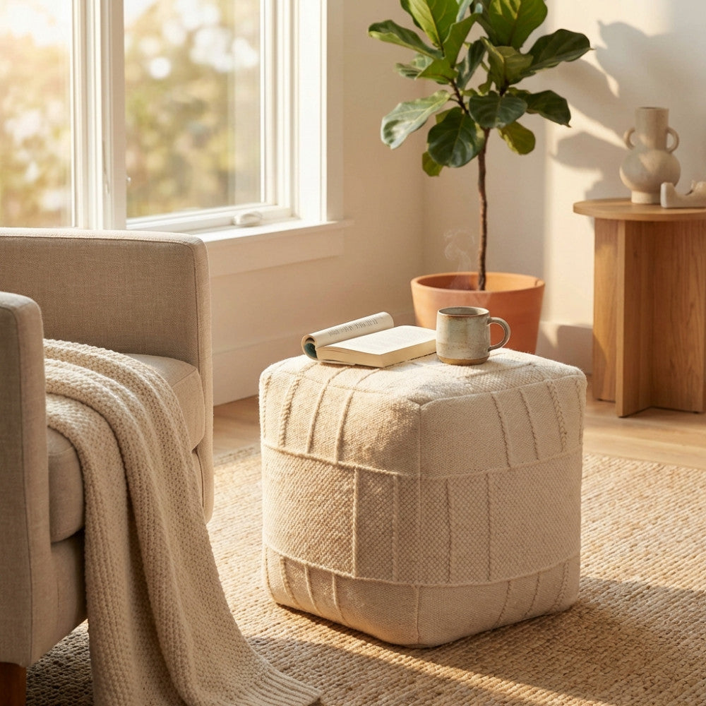 Dettaglio del pouf bianco intrecciato accanto a una poltrona chiara, con una tazza fumante e un libro appoggiati sopra.