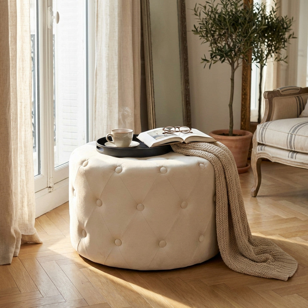 Pouf rotondo in velluto beige posizionato in un salotto luminoso con vassoio e tazza di caffè.