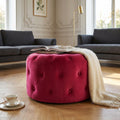 Pouf rotondo in velluto bordeaux posizionato in un salotto elegante con divani scuri, decorato con una coperta chiara e libri.