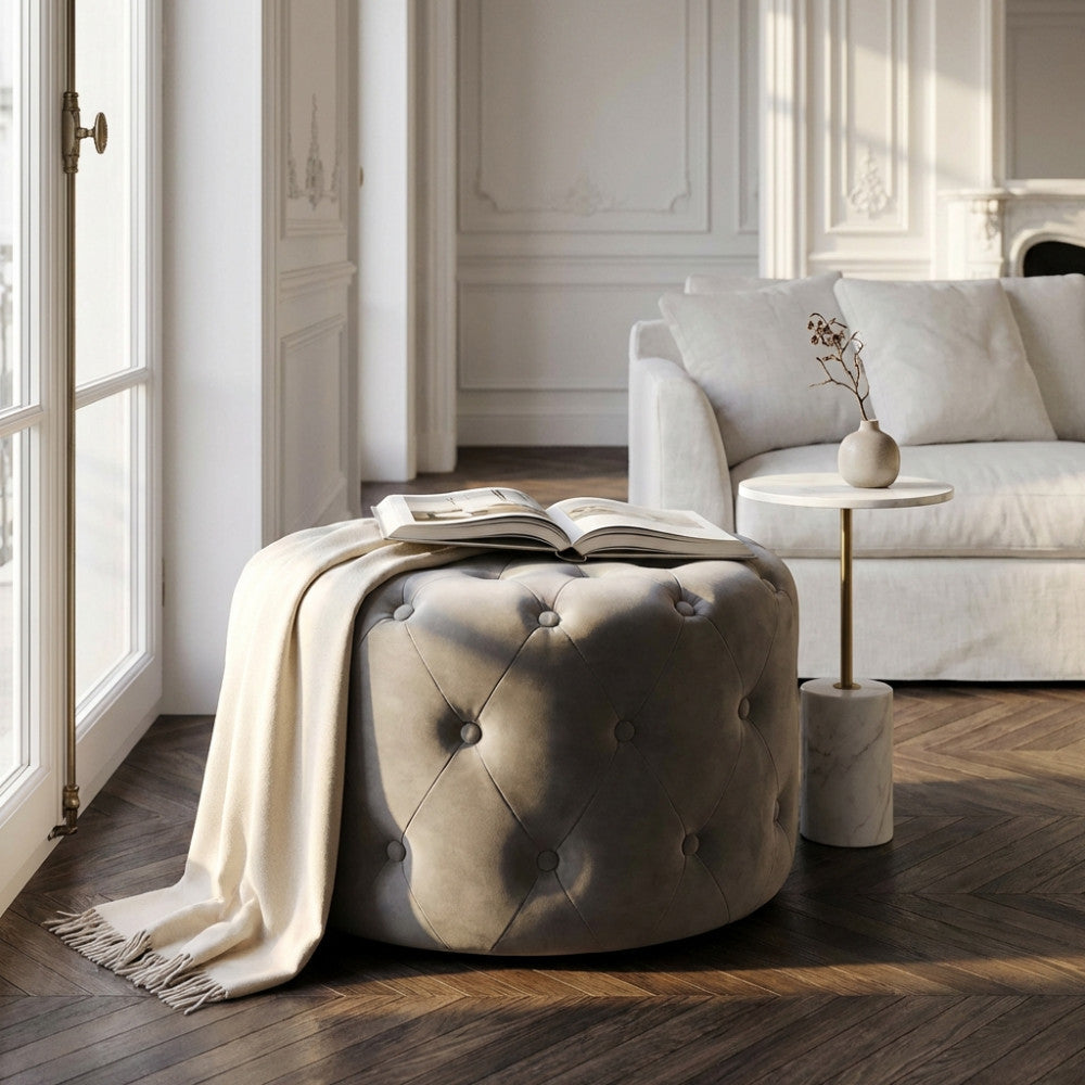 Pouf rotondo in velluto tortora capitonné in un luminoso soggiorno elegante, decorato con un libro e una coperta color panna, posizionato su un pavimento in legno a spina di pesce accanto a un tavolino in marmo.