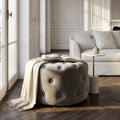 Pouf rotondo in velluto tortora capitonné in un luminoso soggiorno elegante, decorato con un libro e una coperta color panna, posizionato su un pavimento in legno a spina di pesce accanto a un tavolino in marmo.