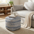 Pouf Sébastien in lana e cotone blu e bianco posizionato su un tappeto chiaro in un salotto moderno, utilizzato come piano d'appoggio per libri e una tazza.