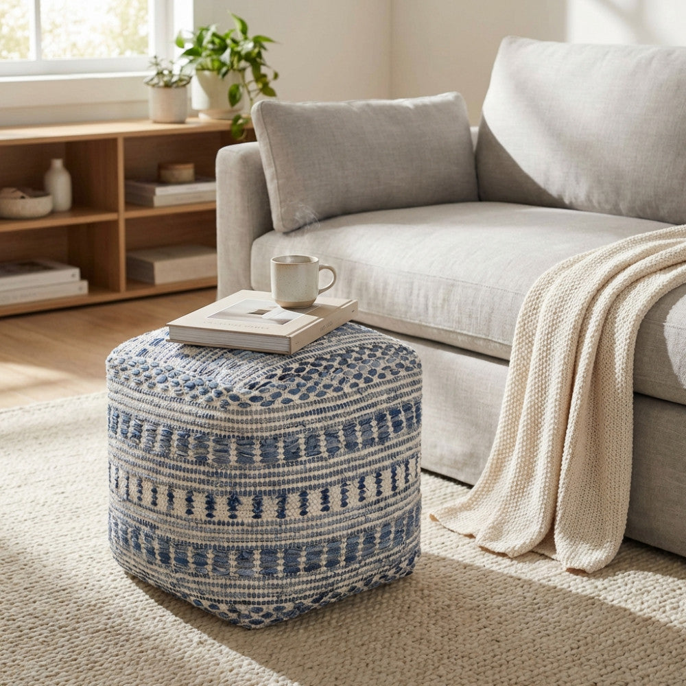 Pouf Sébastien in lana e cotone blu e bianco posizionato su un tappeto chiaro in un salotto moderno, utilizzato come piano d'appoggio per libri e una tazza.