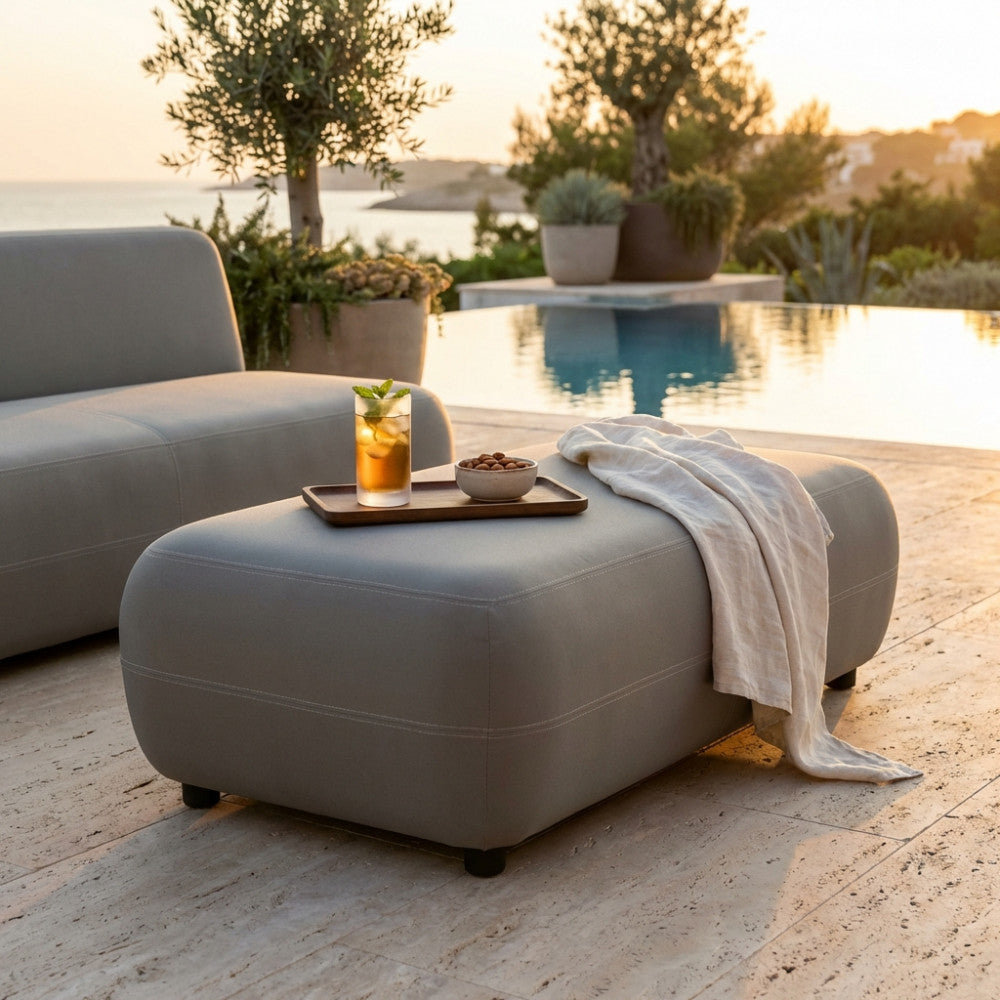 Pouf modulare Tucumán grigio scuro ambientato a bordo piscina al tramonto con vassoio e drink.