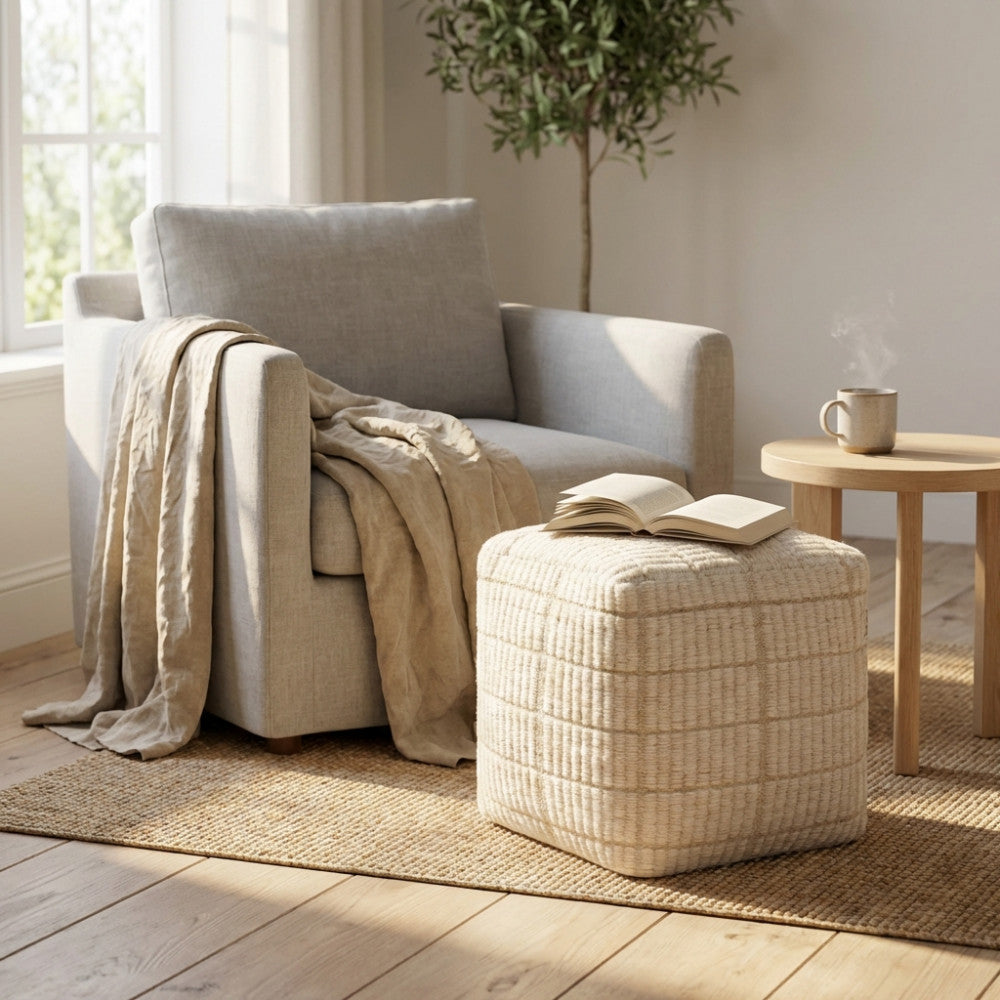 Pouf quadrato in lana intrecciata posizionato vicino a una poltrona in lino e un tavolino in legno.
