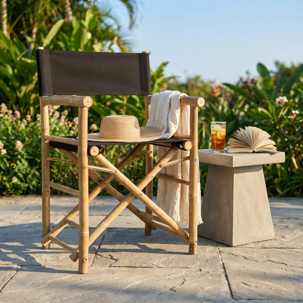 Sedia regista in bamboo con seduta nera, decorata con cappello di paglia e telo chiaro in un giardino lussureggiante.
