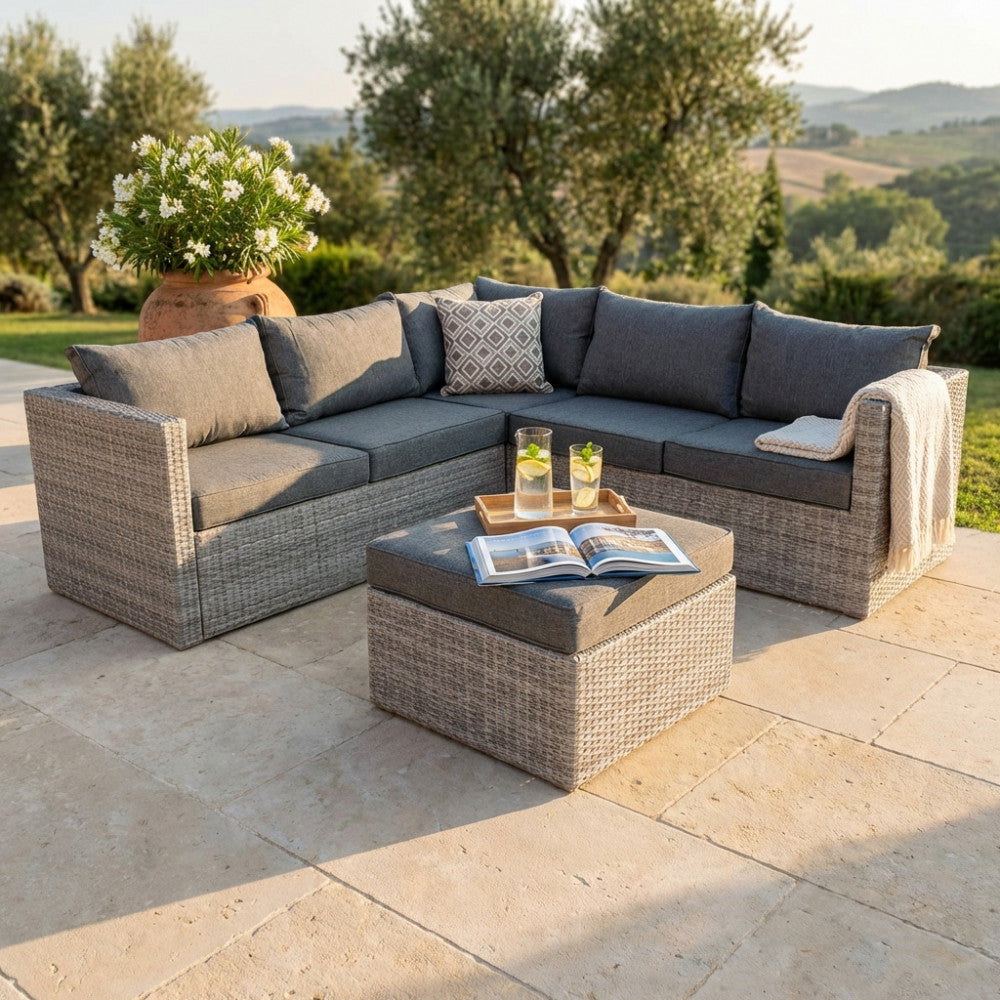 Salotto angolare in wicker grigio con cuscini sfoderabili ambientato in un giardino lussuoso al tramonto.