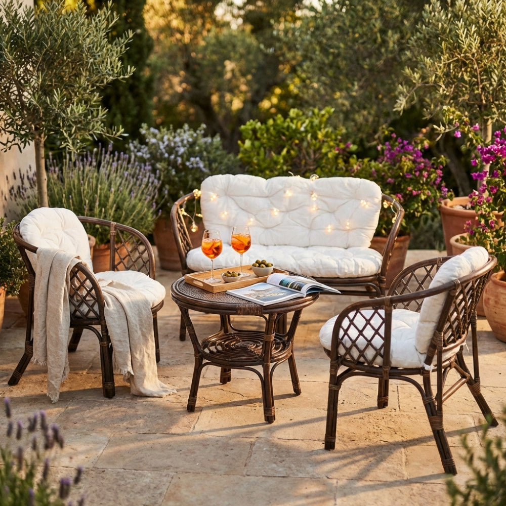 Salotto Bahamas in midollino marrone allestito in un rigoglioso giardino al tramonto, con cuscini in cotone ecru e luci decorative.