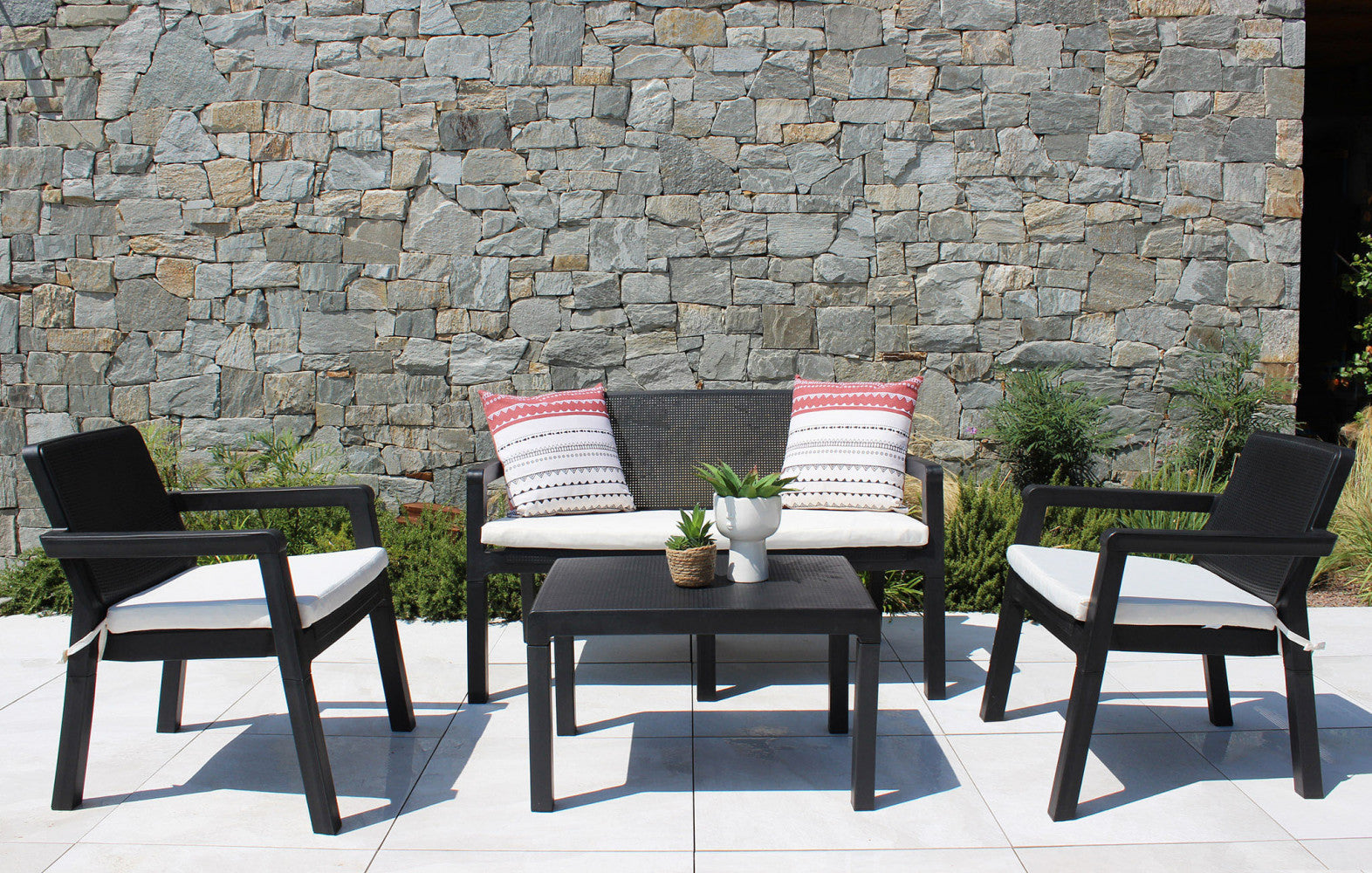 Salotto Delaware nero allestito su una terrazza con pavimentazione chiara e un muro in pietra grezza, arricchito da cuscini decorativi.