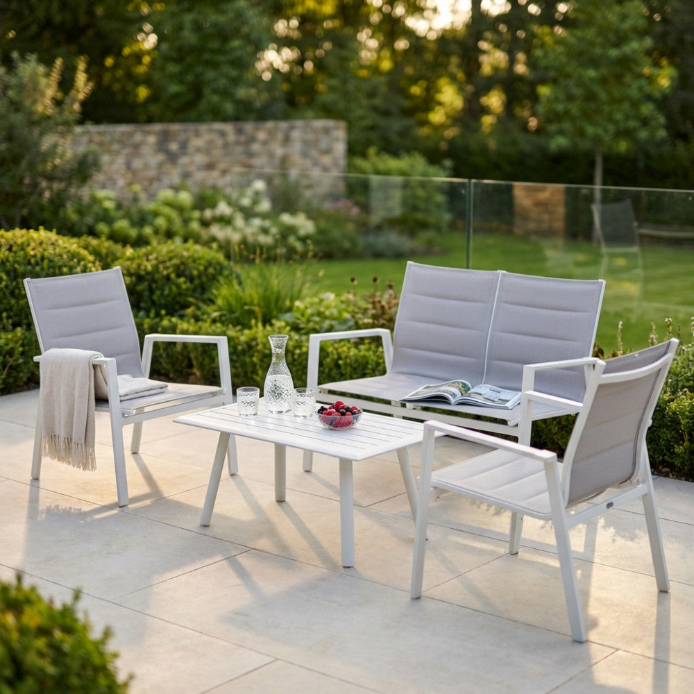 Salotto da esterno Illinois in alluminio bianco e textilene grigio, allestito su un patio in pietra con bicchieri e caraffa sul tavolino, circondato da un giardino curato.