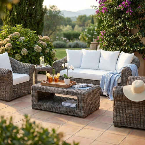 Salotto Karakol in rattan marrone allestito su un patio soleggiato con cuscini bianchi e tavolino centrale.