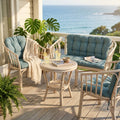 Salotto Turks in rattan bianco con cuscini azzurri allestito su un balcone in legno con vista sull'oceano.