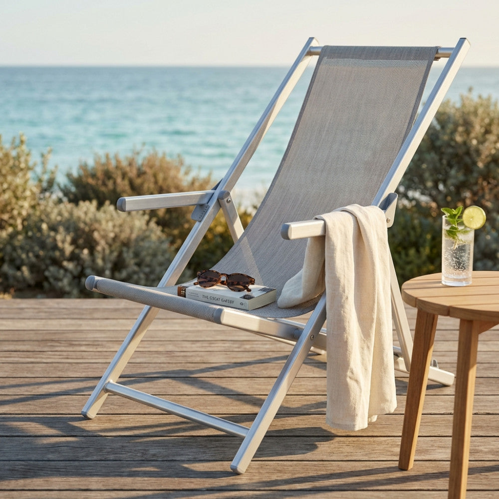 Sdraio pieghevole in alluminio e textilene grigio posizionata su un ponte in legno di fronte al mare, affiancata da un tavolino con drink.