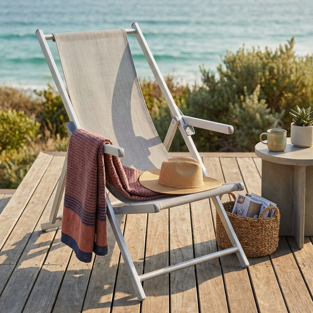 Angolazione laterale della sdraio da giardino grigia su pavimentazione in legno, con accessori estivi e vista sull'oceano.