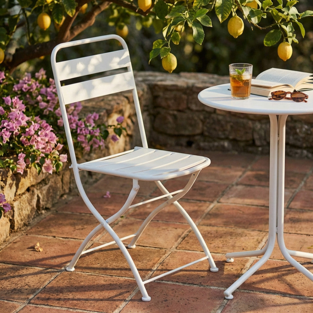 Sedia pieghevole Bristol in acciaio bianco ambientata in un giardino soleggiato accanto a un tavolino.