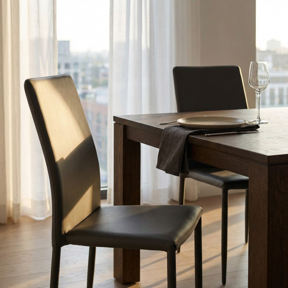 Sedia Cammie in ecopelle grigio scuro posizionata accanto a un tavolo da pranzo in legno scuro, illuminata dalla luce naturale.