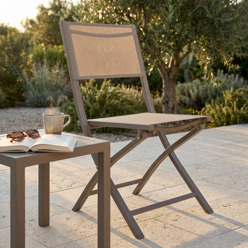 Sedia pieghevole Georgia color taupe in alluminio e textilene, ambientata all'aperto accanto a un tavolino con tazza fumante e libro.
