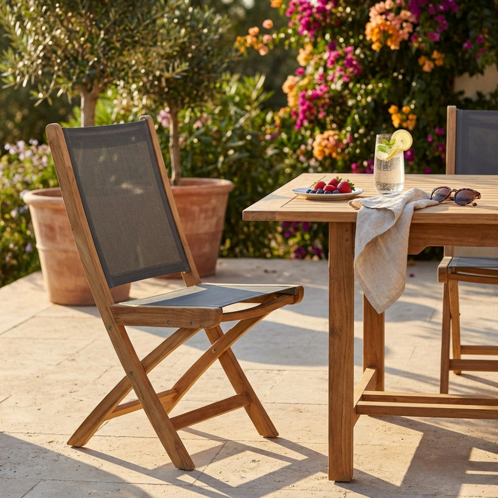 Sedia pieghevole Lahore in teak e textilene grigio posizionata su un patio in pietra accanto a un tavolo da pranzo in legno.