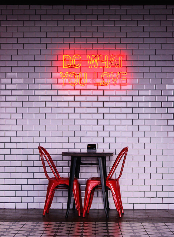 Due sedie Lola Red Antico in acciaio attorno a un tavolino nero, contro un muro di piastrelle bianche con insegna al neon.