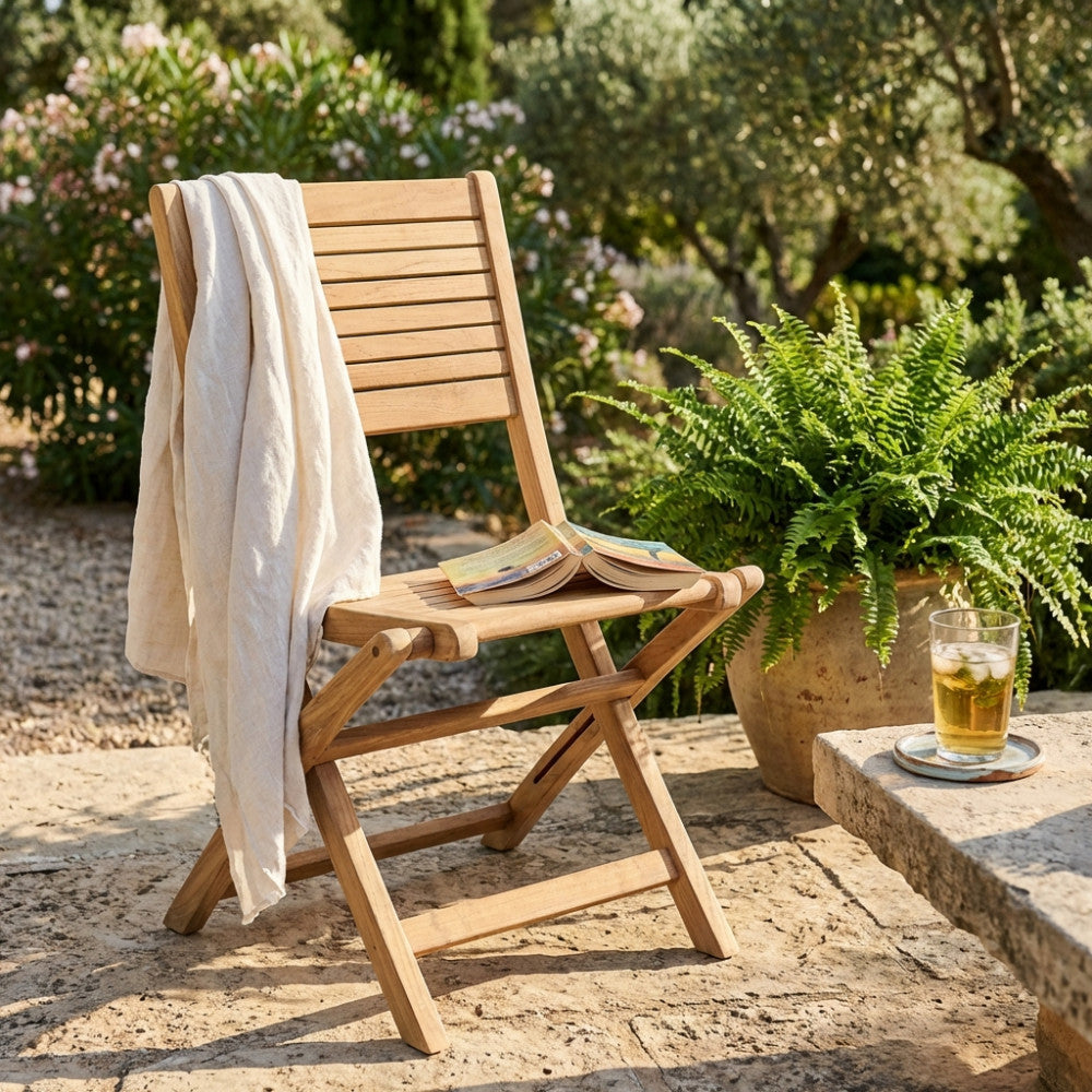 Sedia Mirage pieghevole in legno teak posizionata in un giardino soleggiato con un telo in lino appoggiato sullo schienale.