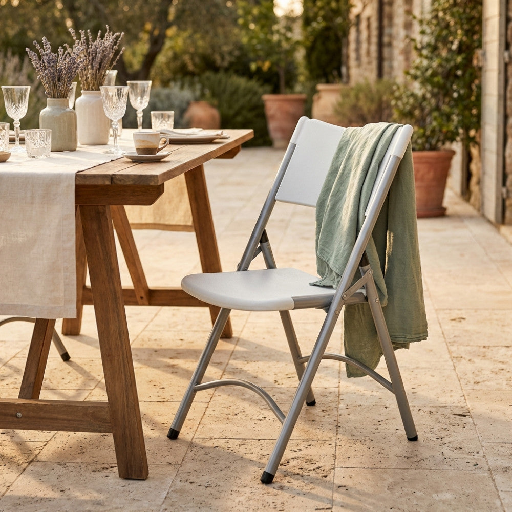 Sedia pieghevole Catering 1 in acciaio e plastica bianca, posizionata all'aperto accanto a un tavolo in legno rustico apparecchiato.